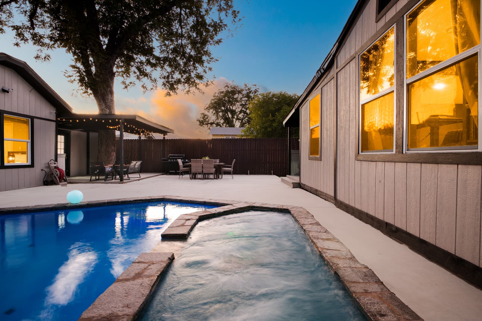 Pool running the length of the lot beneath a mature oak, with the house lit at dusk